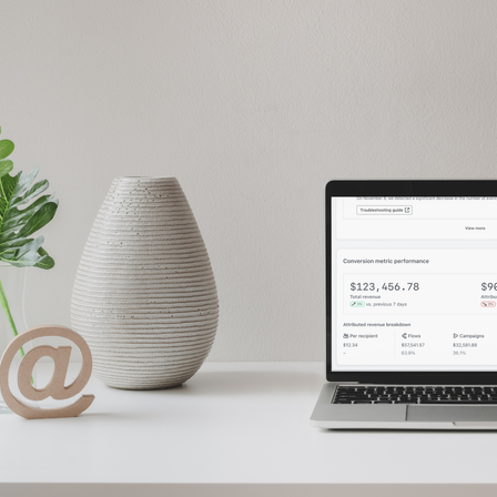A white desk dressed with a white vase, a monstera leaf, a wooden paper weight in the shape of the @ symbol, and a laptop displaying Klaviyo's dashboard