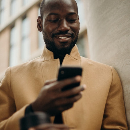 A young man in a camel coat is looking at his phone and smiling.