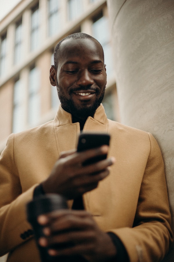 A young man in a camel coat is looking at his phone and smiling.
