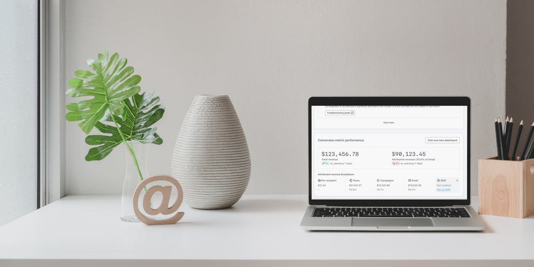 A white desk dressed with a white vase, a monstera leaf, a wooden paper weight in the shape of the @ symbol, and a laptop displaying Klaviyo's dashboard